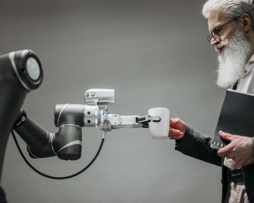 pexels-photo-8439093-8439093 An elderly man receives a cup from a robotic arm in a modern office setting.